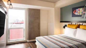 A bedroom with a large bed, a window with blinds, a hanging light fixture, and a wall-mounted TV.
