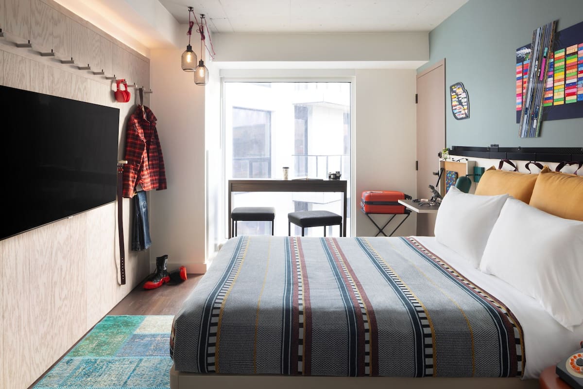 A bedroom with a large bed, a TV mounted on the wall, a balcony with a view, and a desk with a chair.