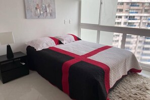 A bedroom with a bed, a nightstand, a lamp, and a window with a view of buildings. - Areia Beach - Pozos Colorados - Santa Marta - Colombia (Santa Marta)