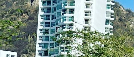 Edificio residencial de varios pisos con balcones, rodeado de vegetación y un paisaje montañoso.
