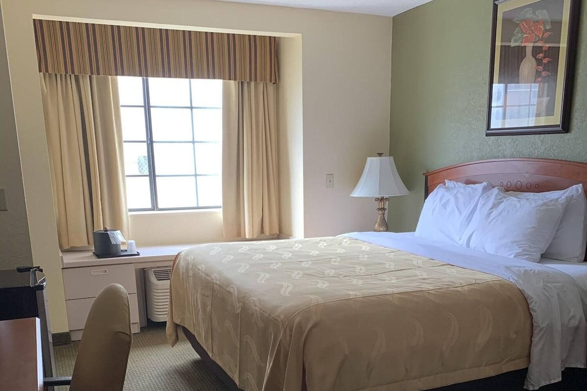 Ein Hotelzimmer mit Bett, Schreibtisch, Stuhl, einem Fenster mit Vorhängen und einem gerahmten Bild an der Wand.