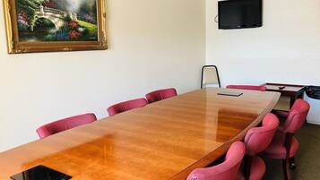 A conference room with a long wooden table, red upholstered chairs, a painting of a bridge, and a flat-screen TV on the wall.