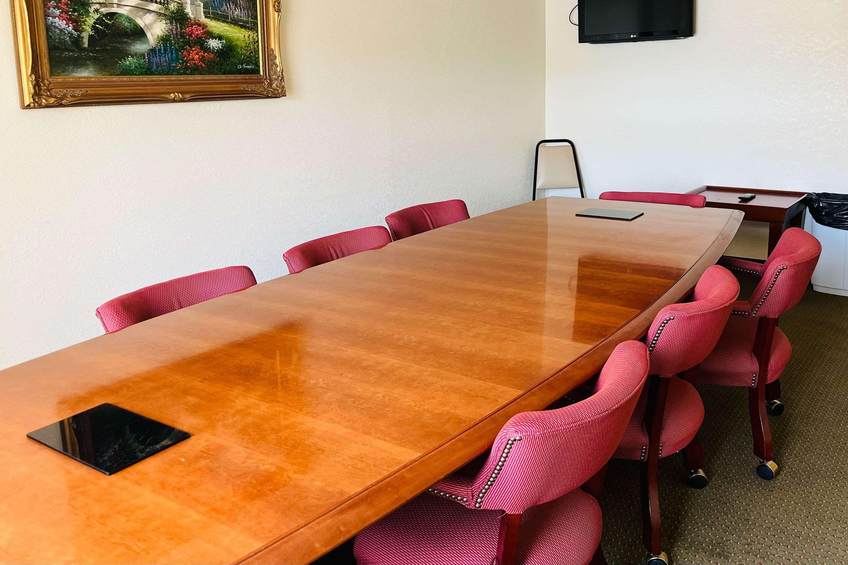 A conference room with a long wooden table, red upholstered chairs, a framed painting, and a television on the wall.