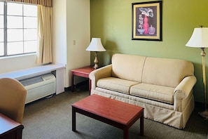 Ein Zimmer mit einem beigen Sofa, einem Holztisch, einem kleinen Tisch mit einer Lampe, einem Heizkörper und einem Fenster mit Vorhängen.