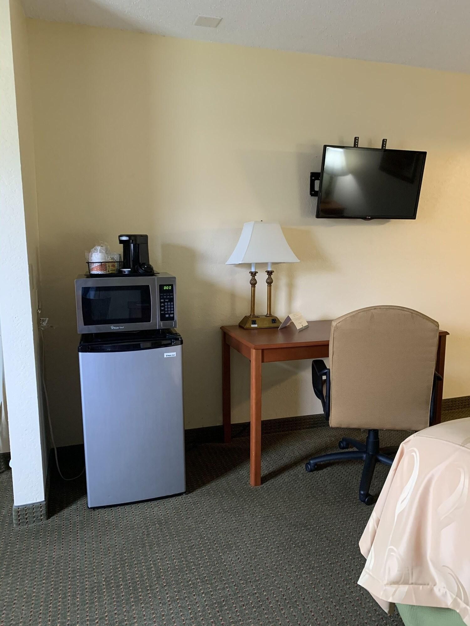 A hotel room with a refrigerator, microwave, small table with a lamp, a TV mounted on the wall, and a chair.