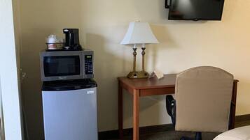 A hotel room with a refrigerator, microwave, small table with a lamp, a TV mounted on the wall, and a chair.