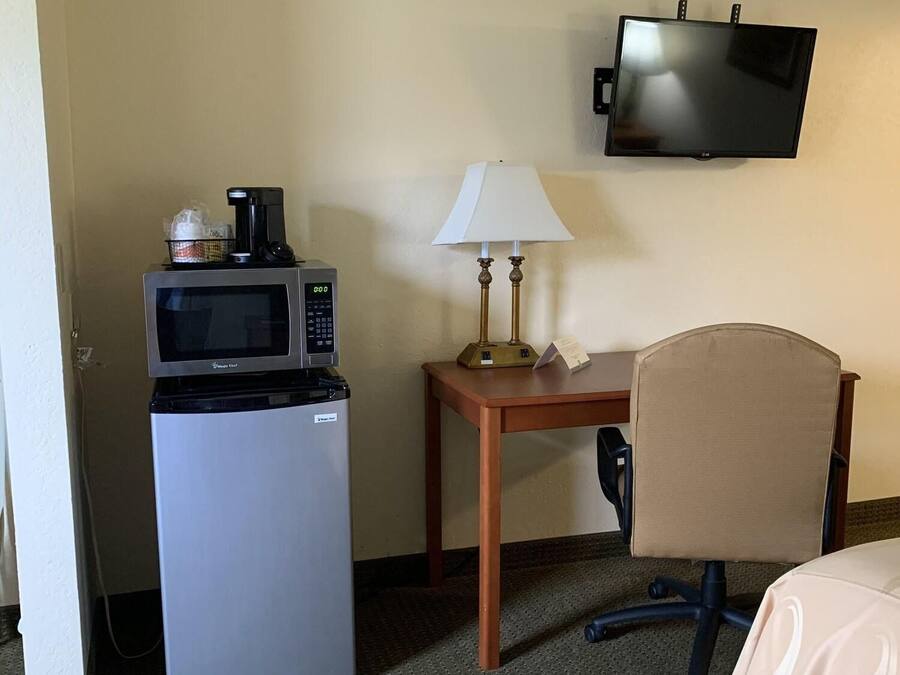 A hotel room with a refrigerator, microwave, small table with a lamp, a TV mounted on the wall, and a chair.