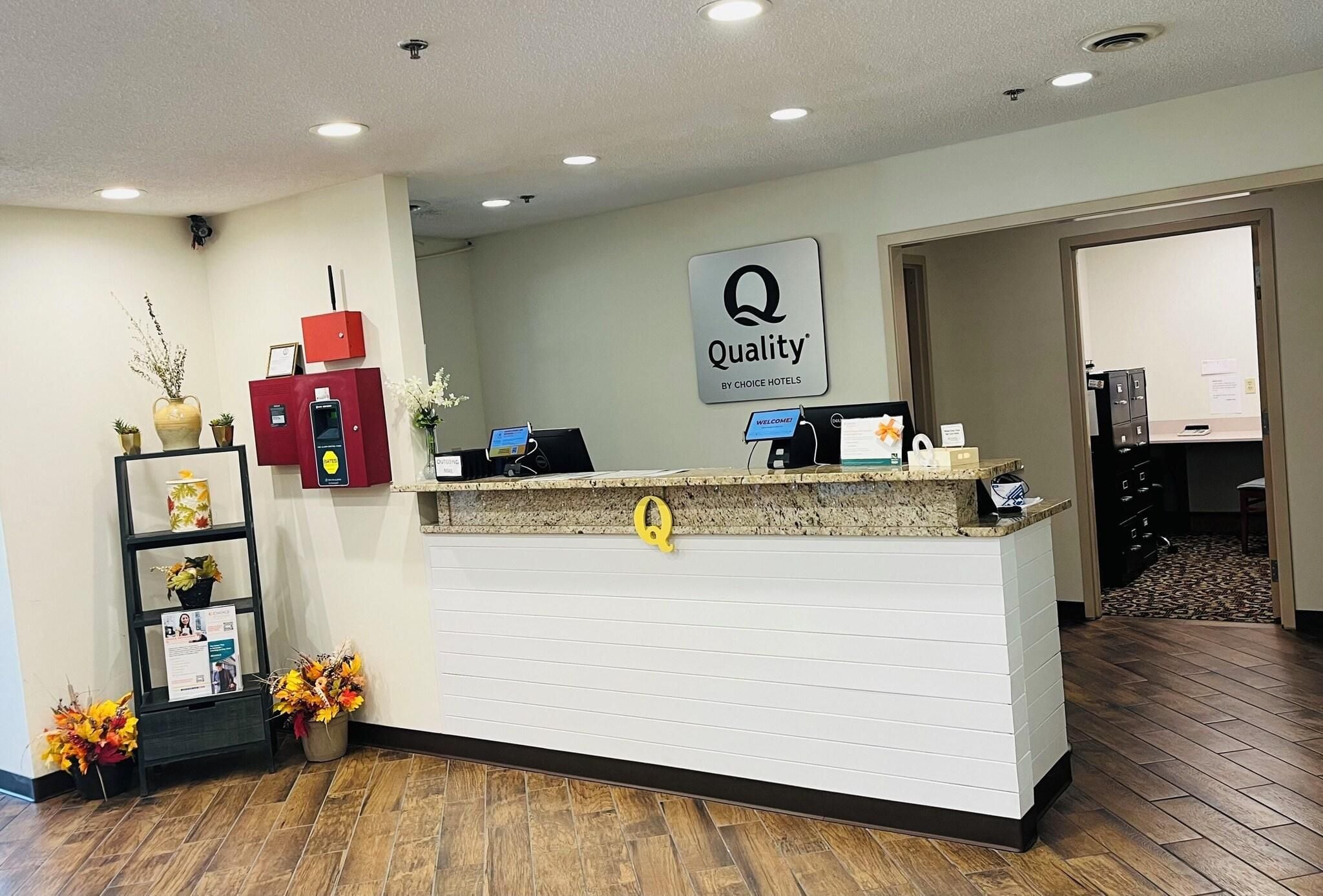 Una recepción de hotel con un cartel de 'Quality by Choice Hotels', un quiosco informativo rojo y un mostrador de recepción.