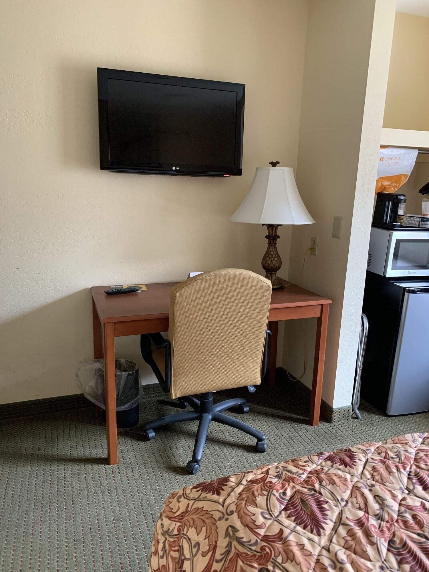 A hotel room with a bed, a desk with a chair, a television, a lamp, and a kitchenette.
