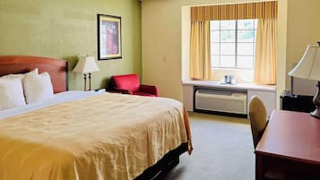 A hotel room with a bed, a desk, a chair, a window with curtains, and a wall-mounted air conditioner.