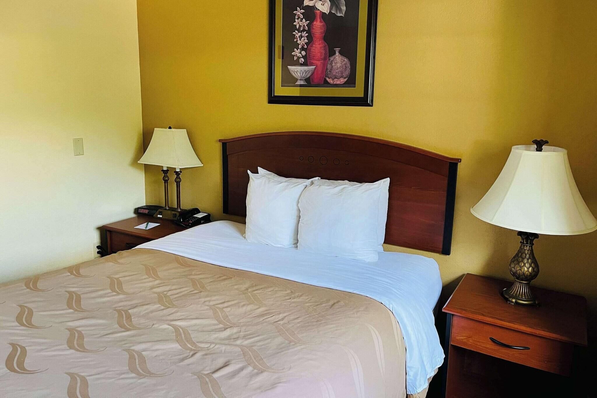 Une chambre d’hôtel avec un lit, deux tables de chevet avec des lampes, une petite table avec un téléphone et un tableau au mur.