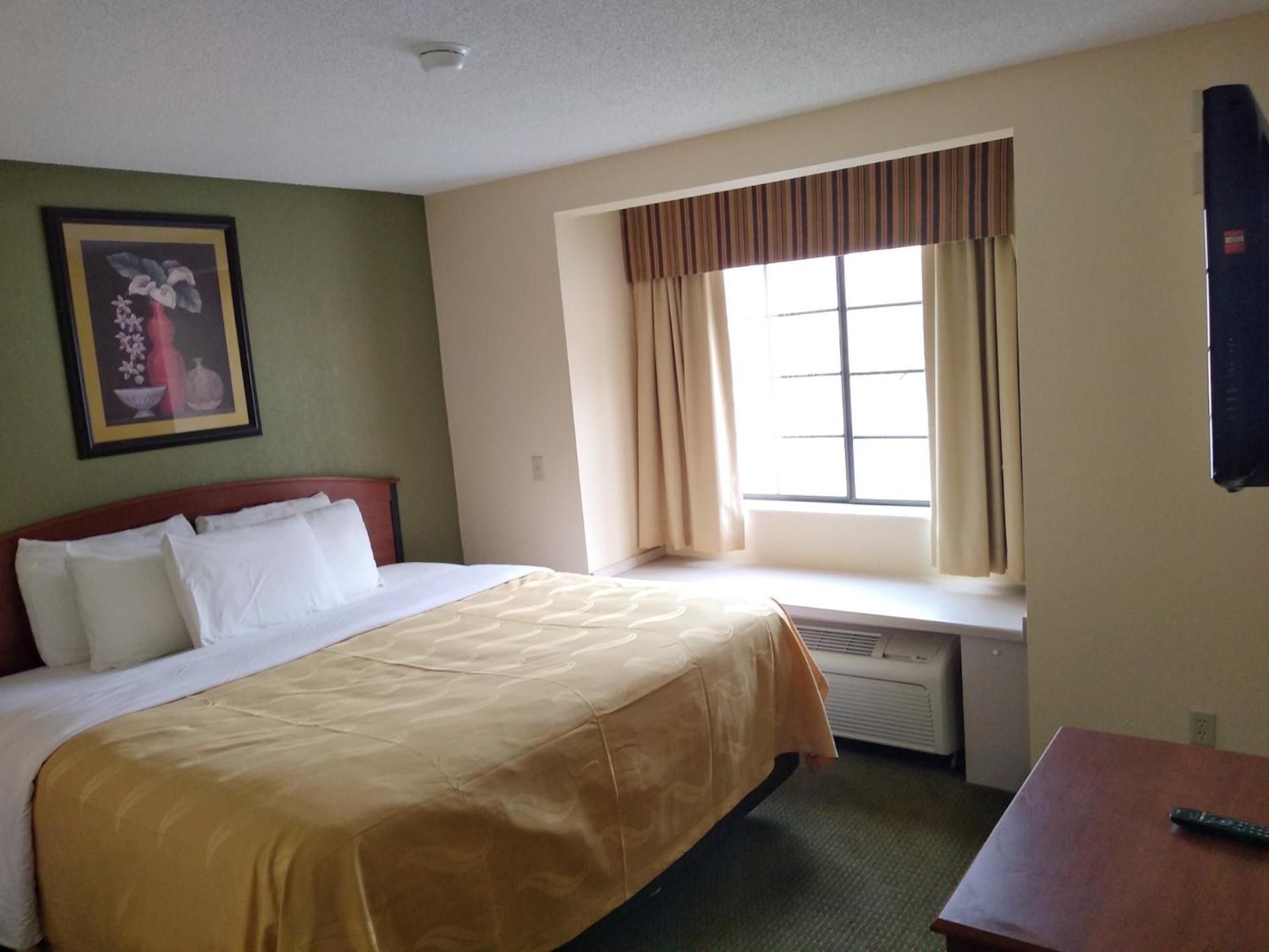 A hotel room with a bed, a window with curtains, a wall-mounted air conditioner, and a framed picture.
