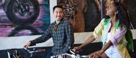 Two people on bicycles with baskets, a mural in the background, and a brick wall.
