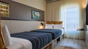Een hotelkamer met twee bedden, een groot raam met gordijnen, een schilderij aan de muur en een tafeltje met een lamp.