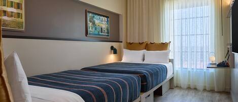 A hotel room with two beds, a large window with curtains, a painting on the wall, and a small table with a lamp.