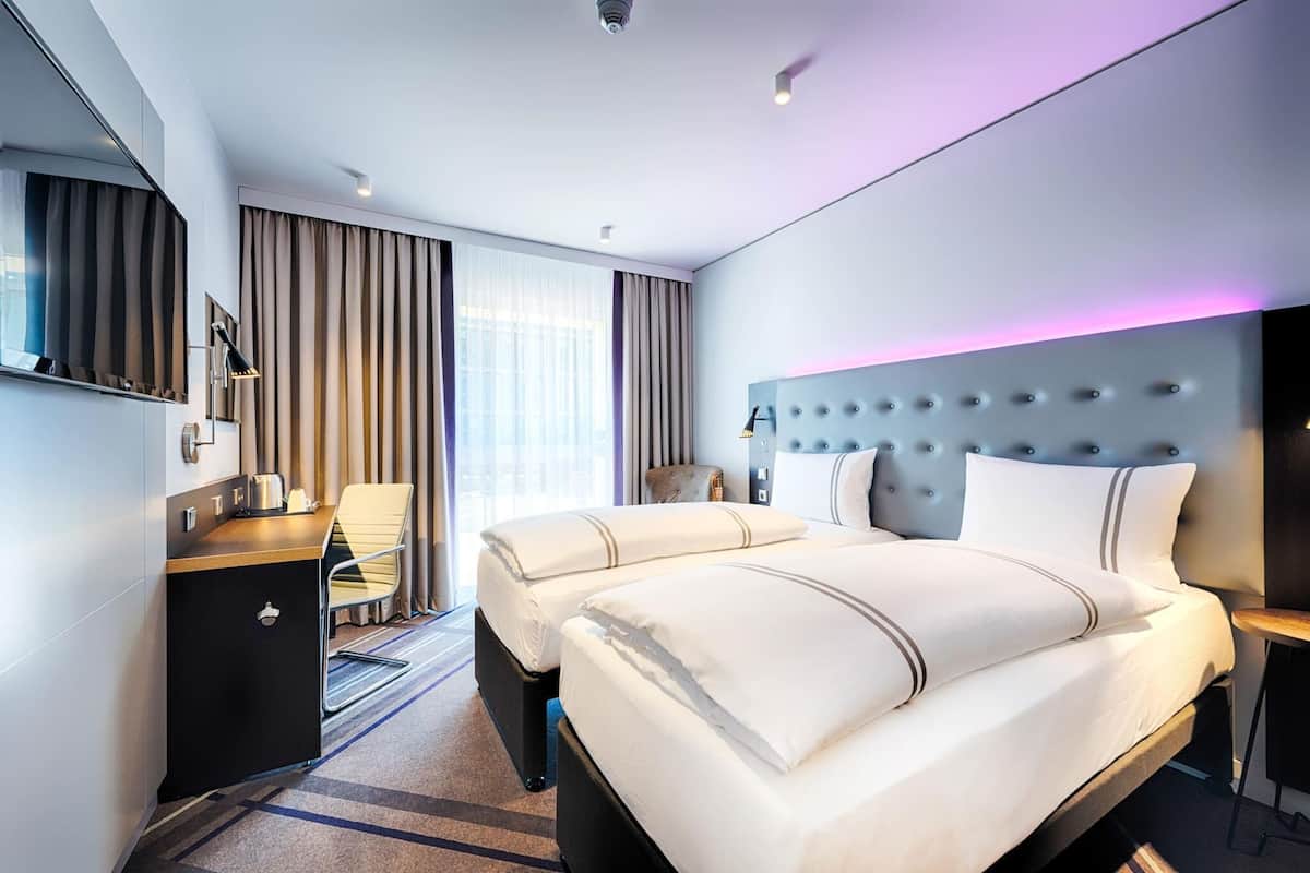 A modern hotel room with a large bed, a desk, a chair, and a minibar.