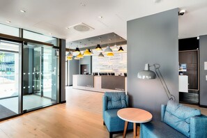 Un moderno vestíbulo de hotel con puertas de vidrio, mostrador de recepción, asientos azules e iluminación colgante.
