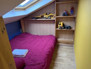 Um quarto com claraboia, uma cama com colcha rosa, prateleiras de madeira com veículos de brinquedo e piso de madeira.