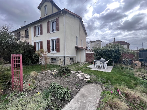 Uma casa de dois andares com jardim, um portão vermelho e uma mesa com cadeiras.