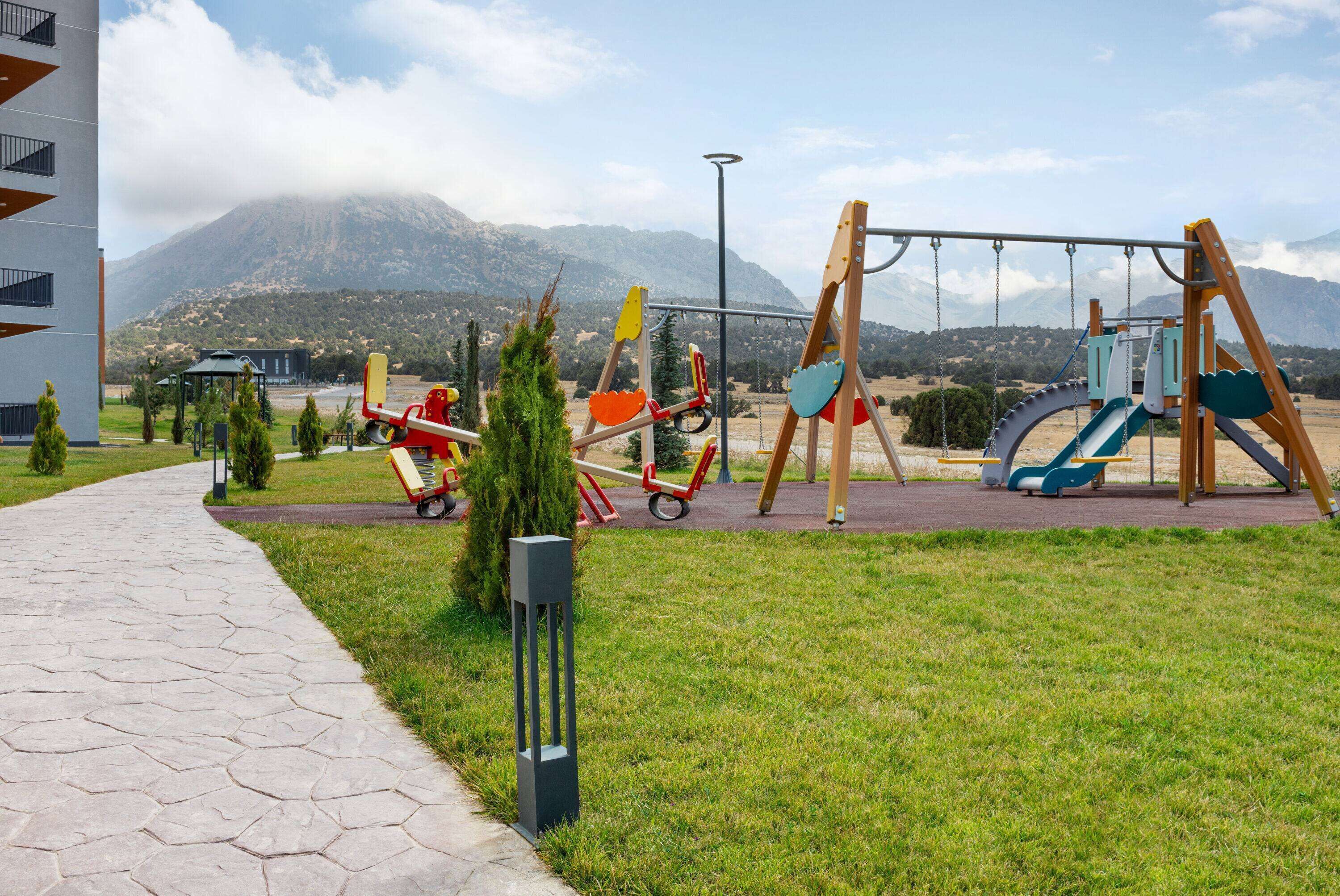Ein Spielplatz mit Schaukeln und Rutschen, ein gepflasterter Weg und ein Berg im Hintergrund.