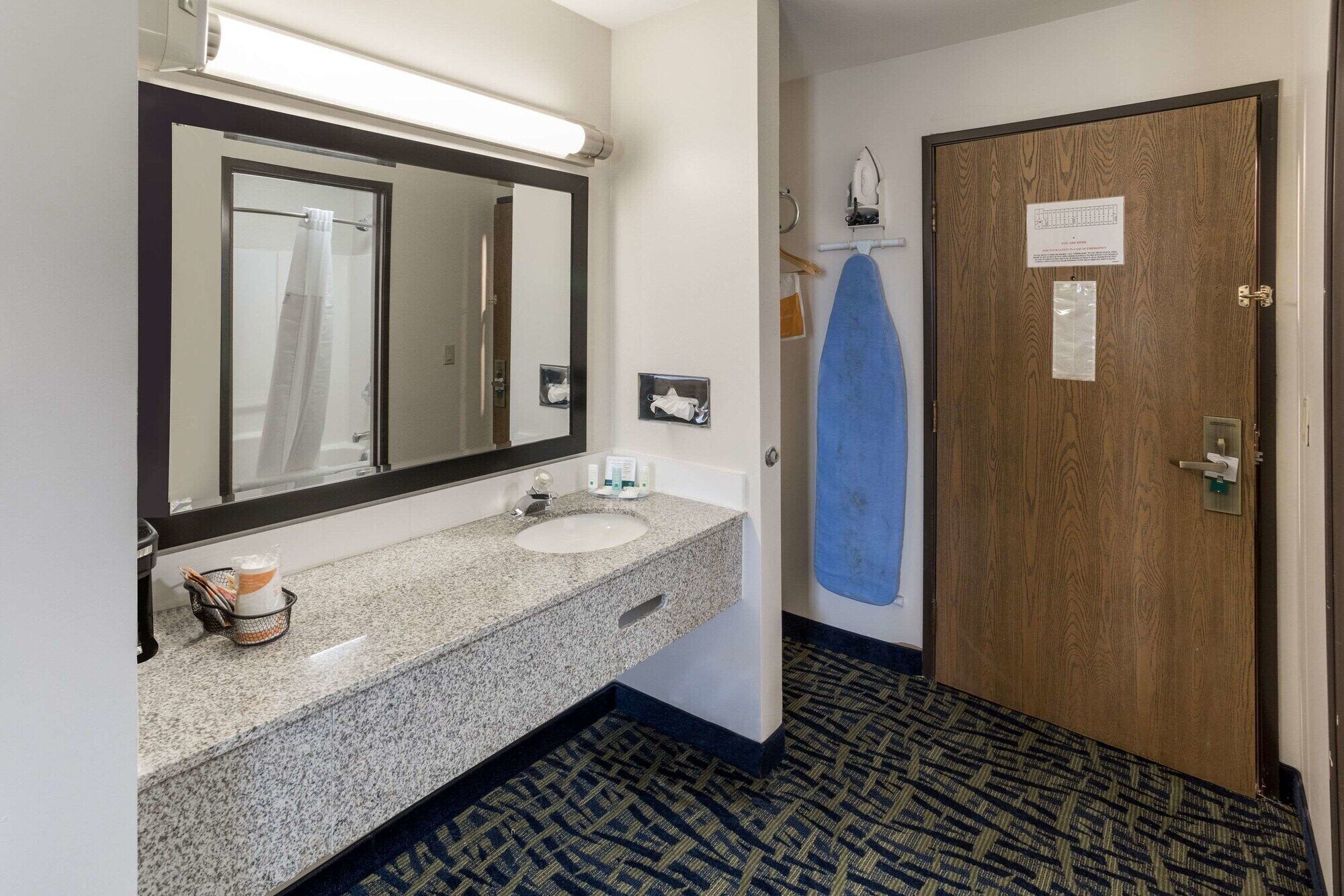 A hotel bathroom with a granite sink, a mirror, a towel rack, and a door with a sign.