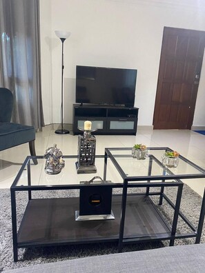 A living room with a glass coffee table, a TV stand, a lamp, and a door.