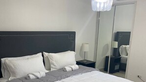 Un lit bien fait, avec une couette grise et blanche, une tête de lit sombre et un lustre moderne.