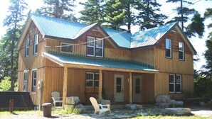 Une cabane en bois à deux étages, avec un toit en métal, une véranda dotée de chaises et un chemin de pierres menant à l’entrée.