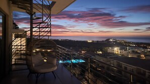 En balkon med stol, pool og udsigt over havet og bybilledet i skumringen.