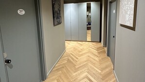 Ein Flur mit Fischgrätparkett, grauen Wänden und einer Decke mit eingelassenen Leuchten. Links und rechts befinden sich Türen, sowie eine Tür am Ende des Flurs.