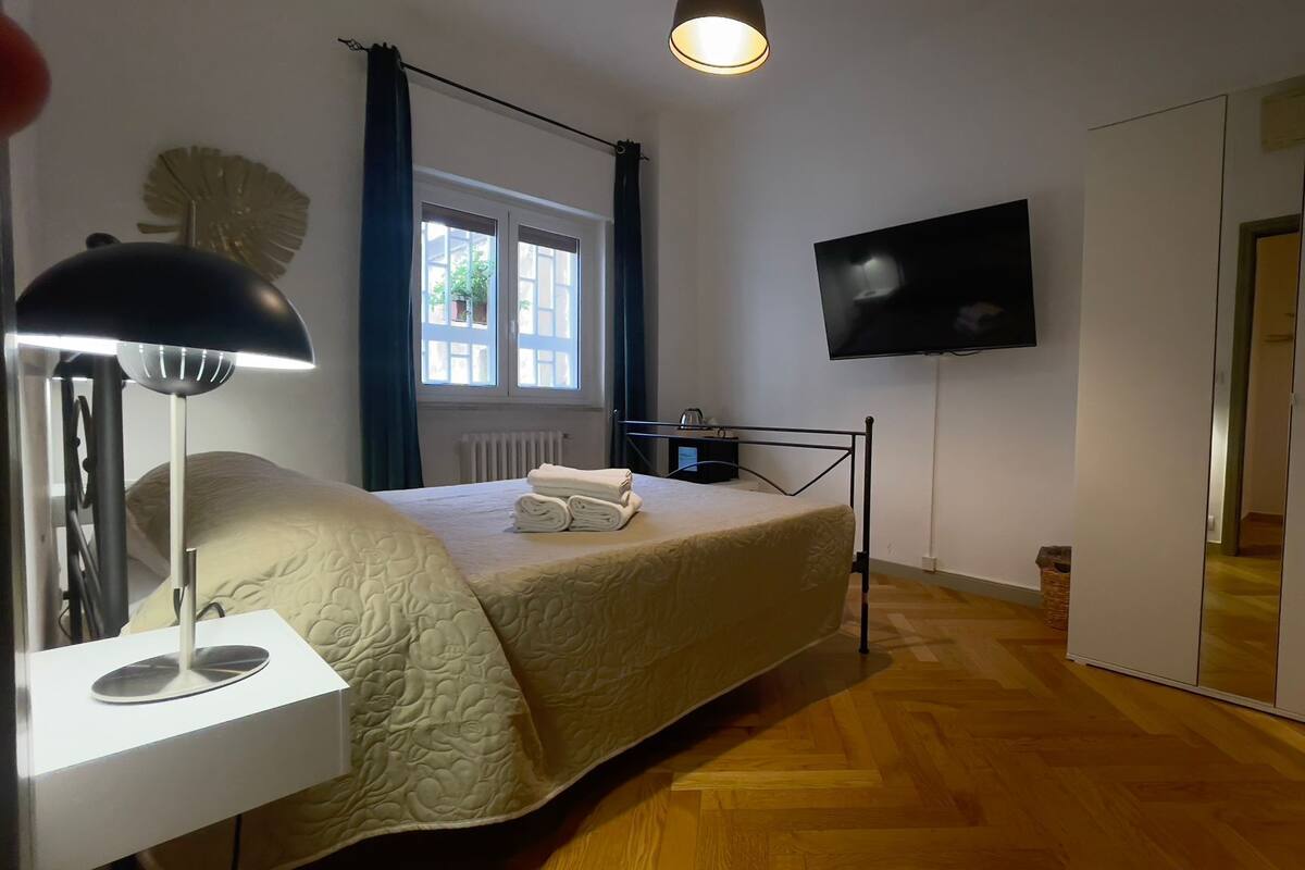 A bedroom with a bed, a bedside lamp, a TV mounted on the wall, and a window with curtains.