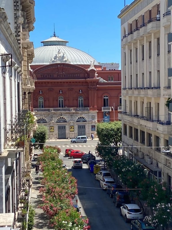 Une vue de rue avec un imposant bâtiment rouge surmonté d’un dôme, sur lequel on peut lire l’inscription « Teatro Petruzzelli ». Des voitures sont garées et quelques piétons se trouvent sur le trottoir.
