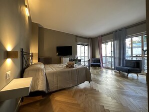 A modern hotel room with a large bed, a flat-screen TV, a sofa, and a view of the city through large windows. - Suite&Chic (Bari)