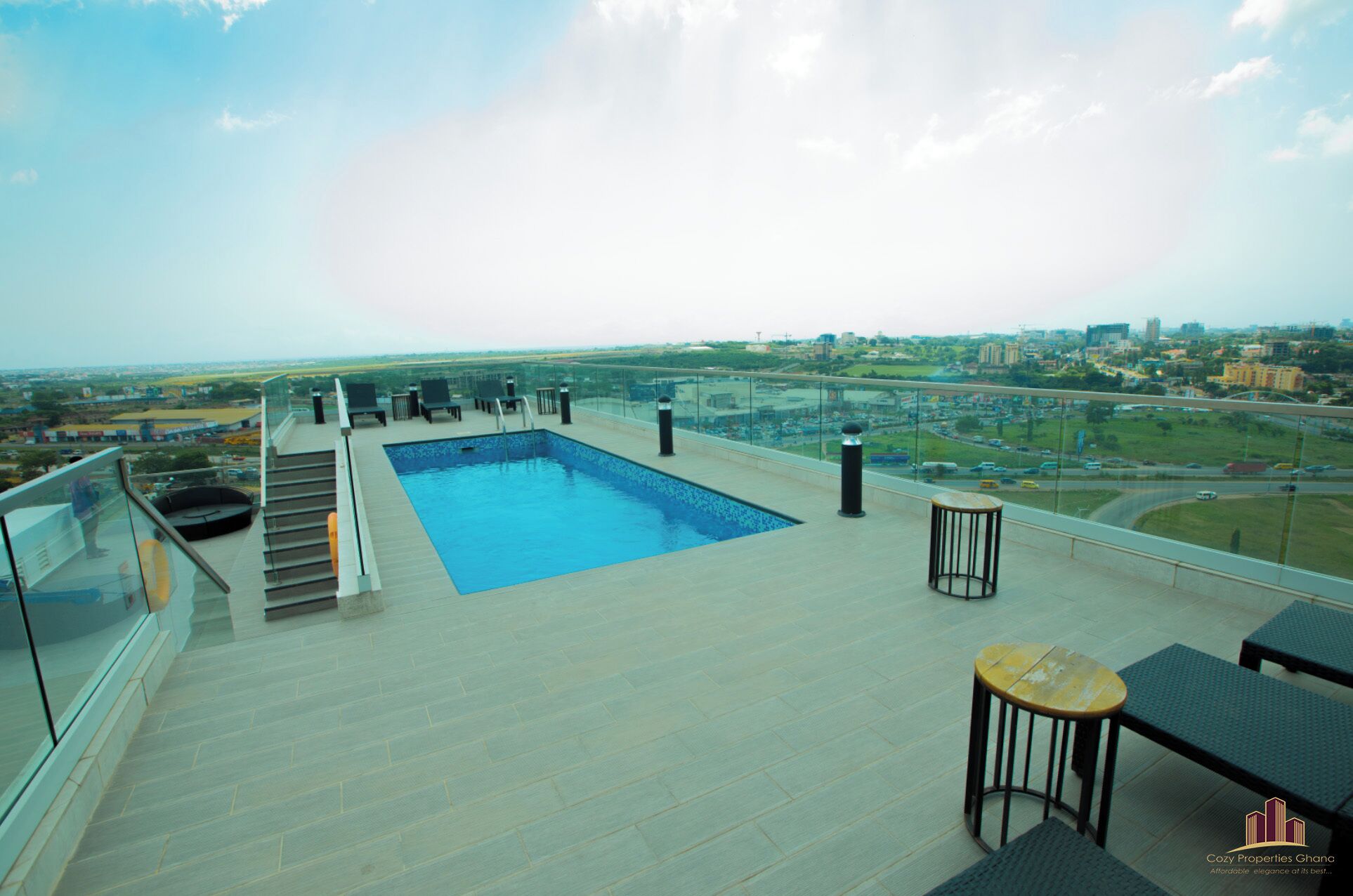Una piscina en la azotea con vistas a la ciudad, barandilla de cristal y mobiliario exterior.