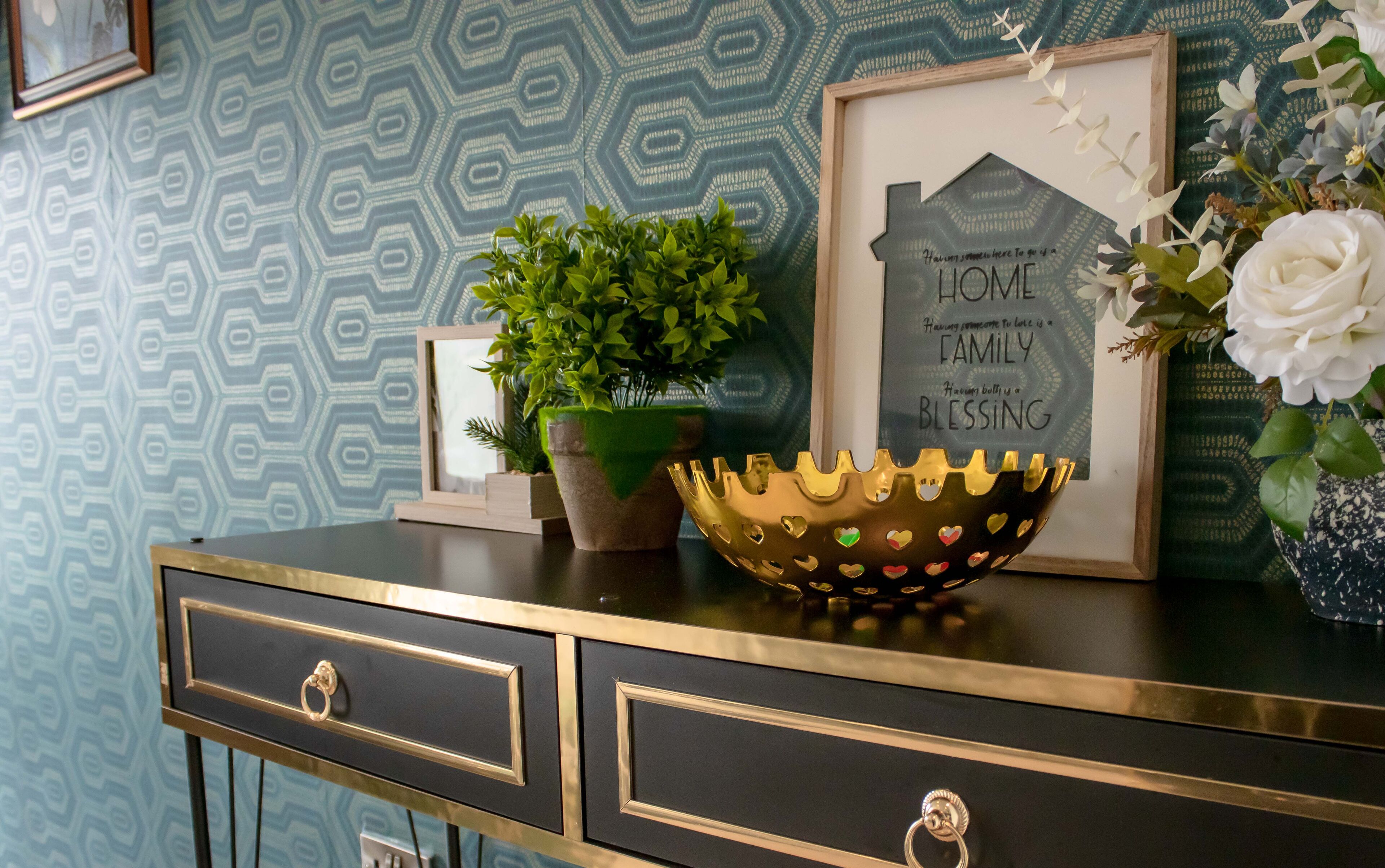 A decorative bowl with heart-shaped cutouts, a framed inspirational quote, a potted plant, and a vase with white flowers on a black and gold console table.
