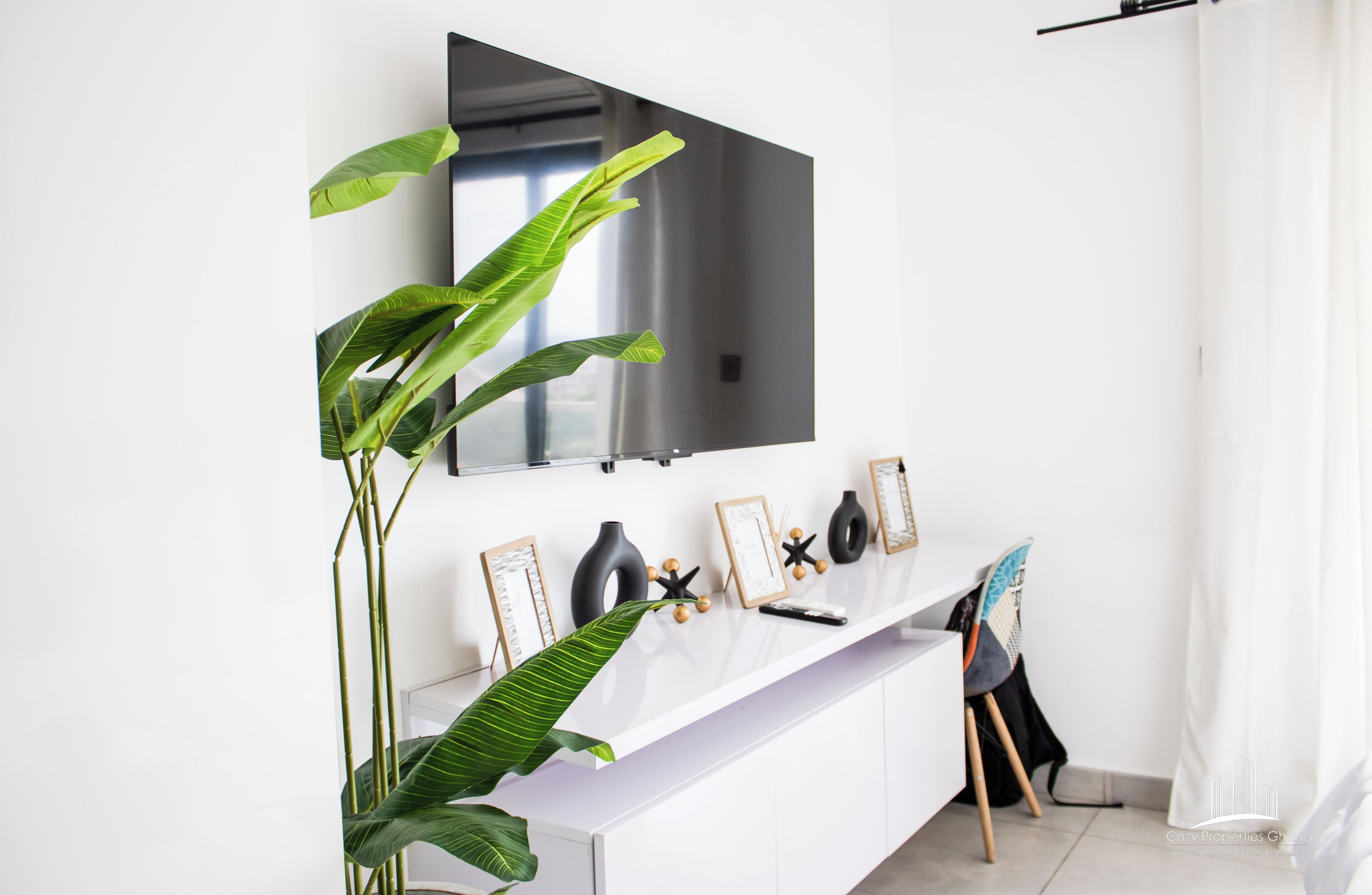 A modern living room with a white console table, a large flat-screen TV mounted on the wall, and a potted plant.