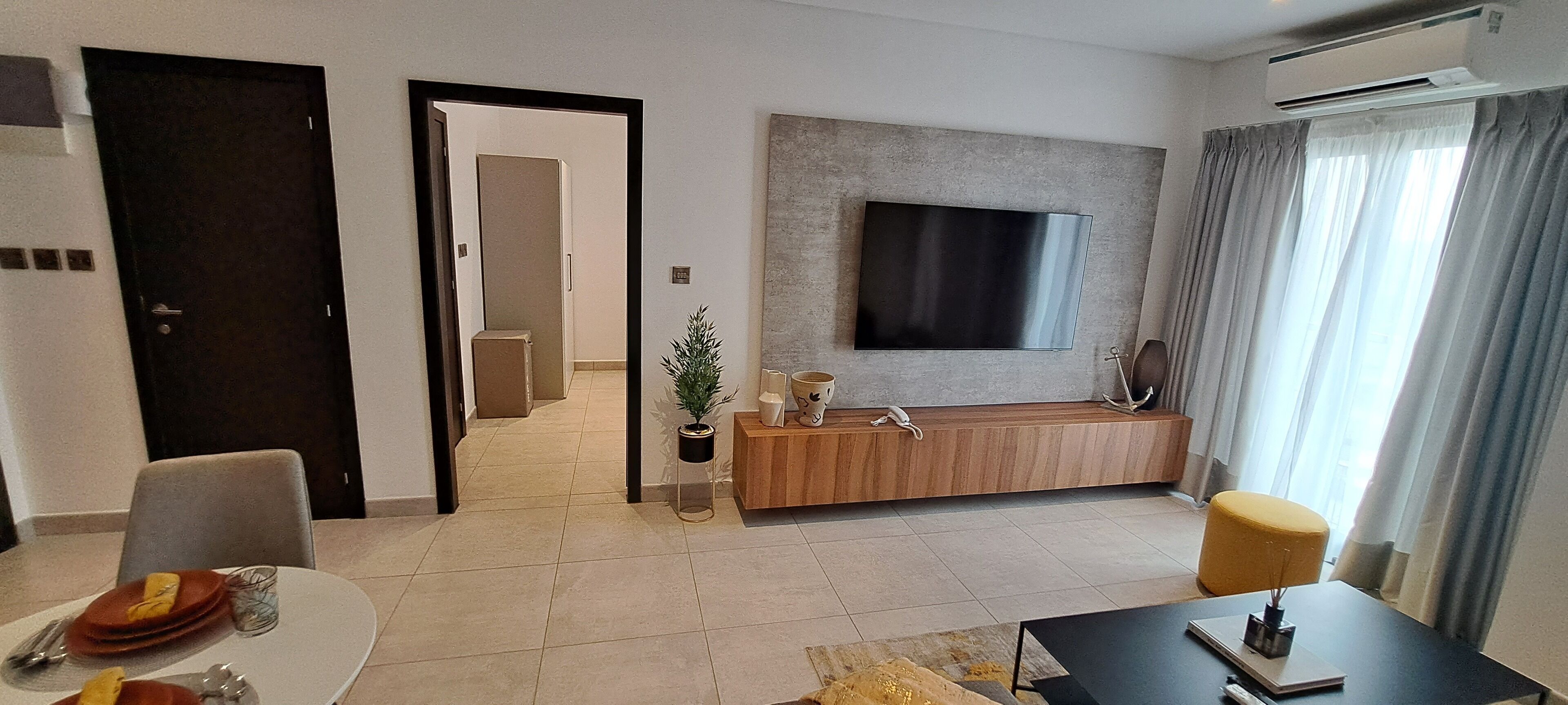 A modern living room with a flat-screen TV mounted on the wall, a wooden console table, and a dining area with a table set for two.