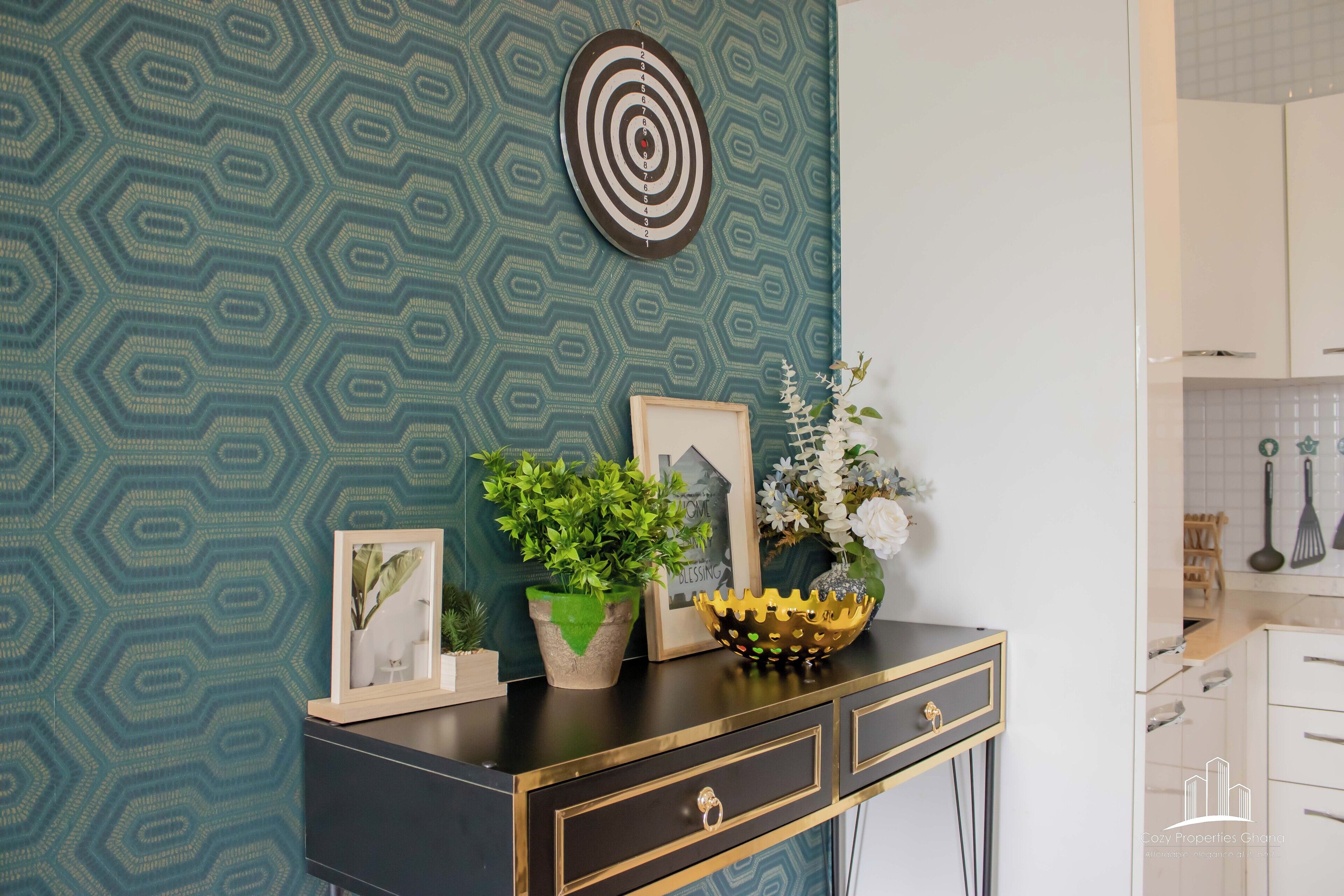 A kitchen with a dartboard, framed pictures, a potted plant, and a decorative bowl on a black and gold console.