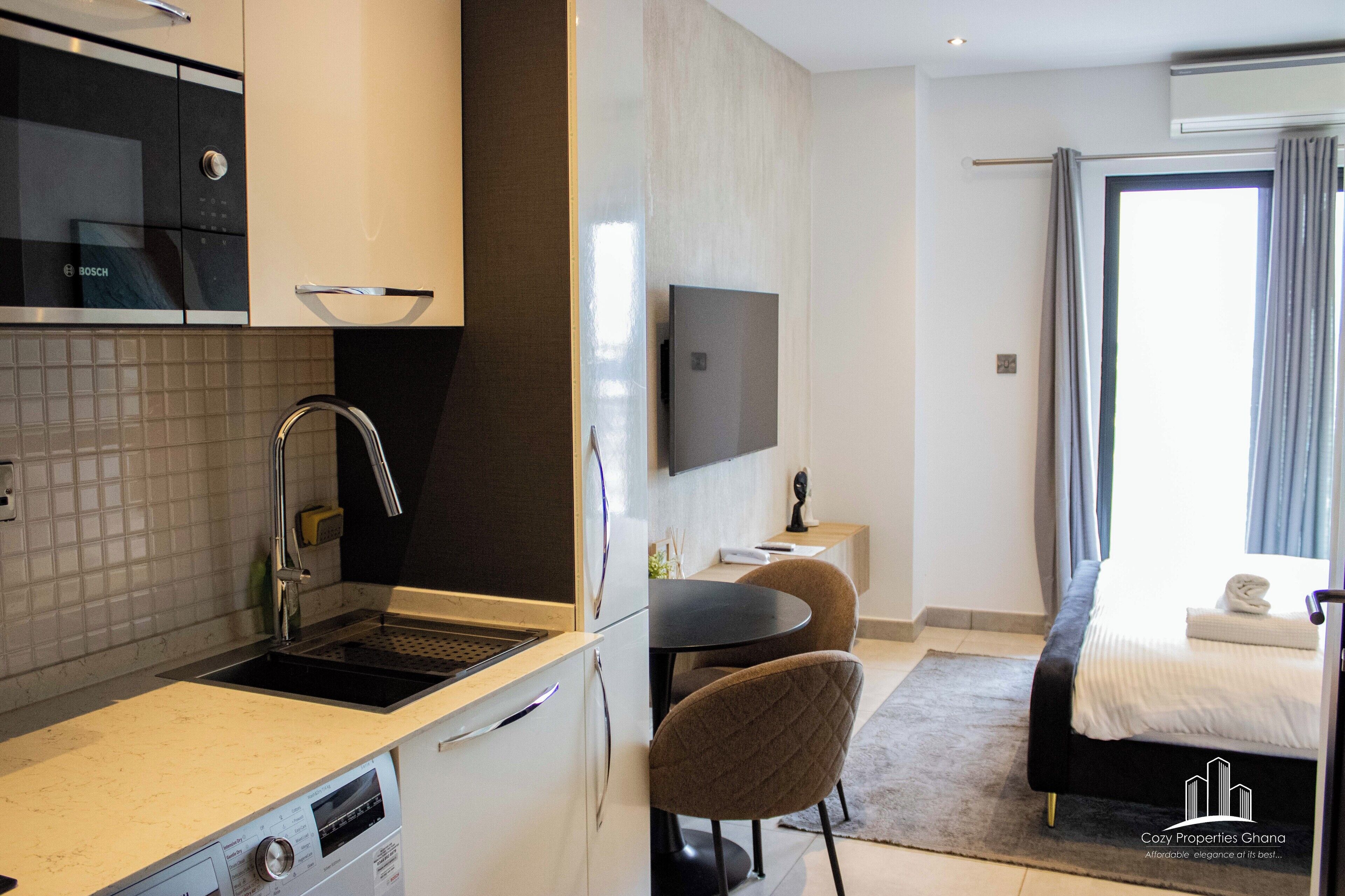 A modern hotel room with a kitchen area, a dining table with chairs, a bed with a view, and a flat-screen TV.
