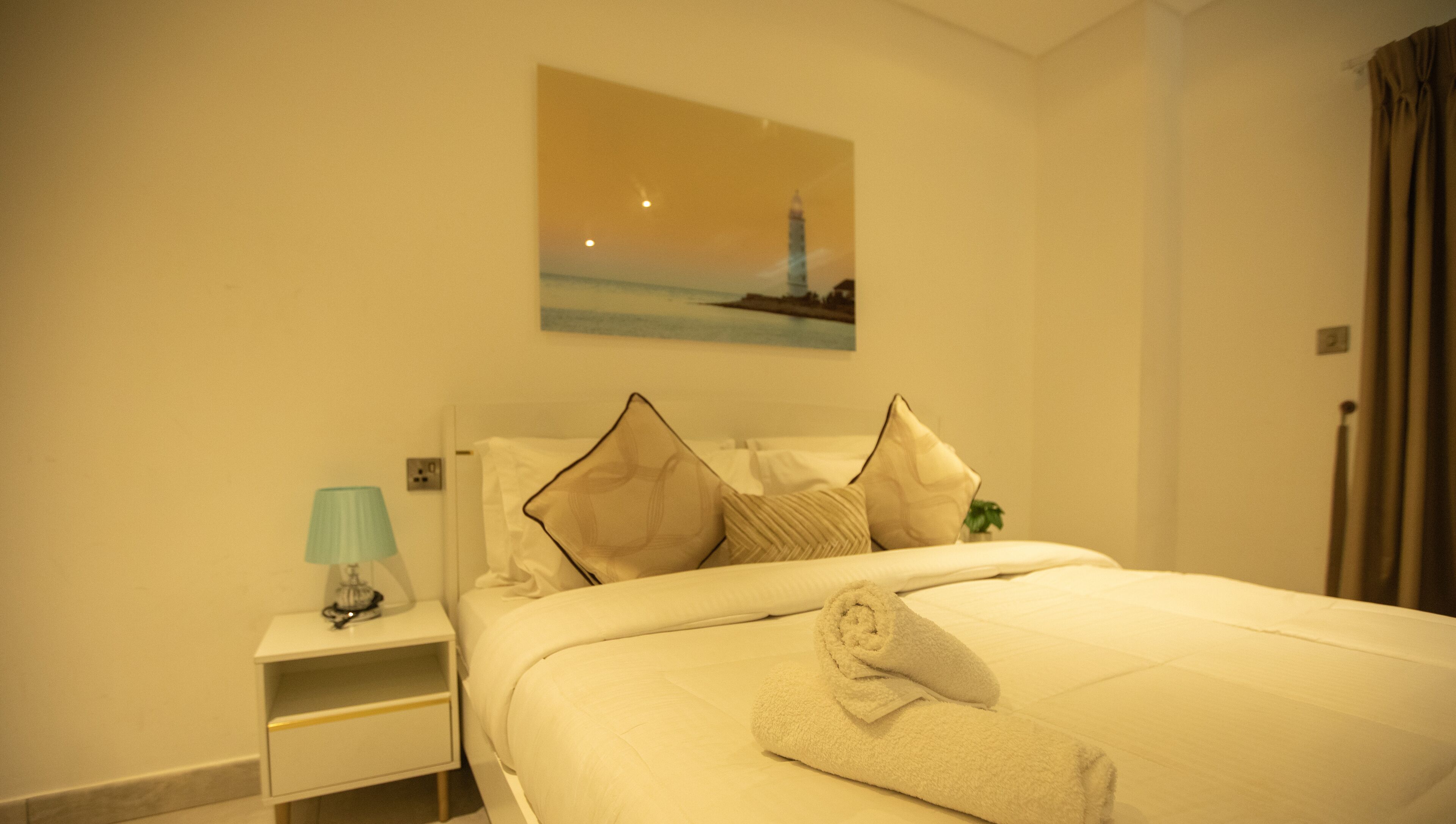 A hotel room with a bed, a nightstand, a lamp, and a painting of a lighthouse on the wall.