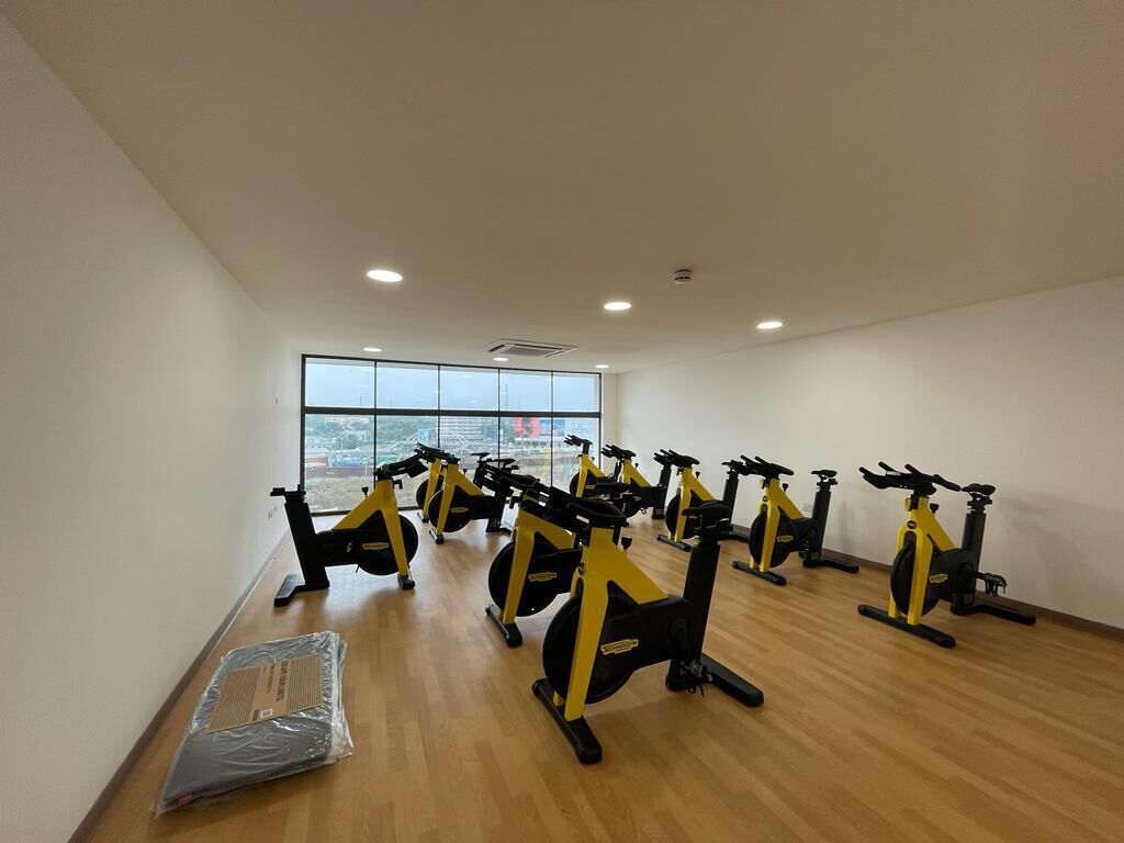 A room with multiple stationary bikes arranged in rows, a large window with a view, and a wooden floor.
