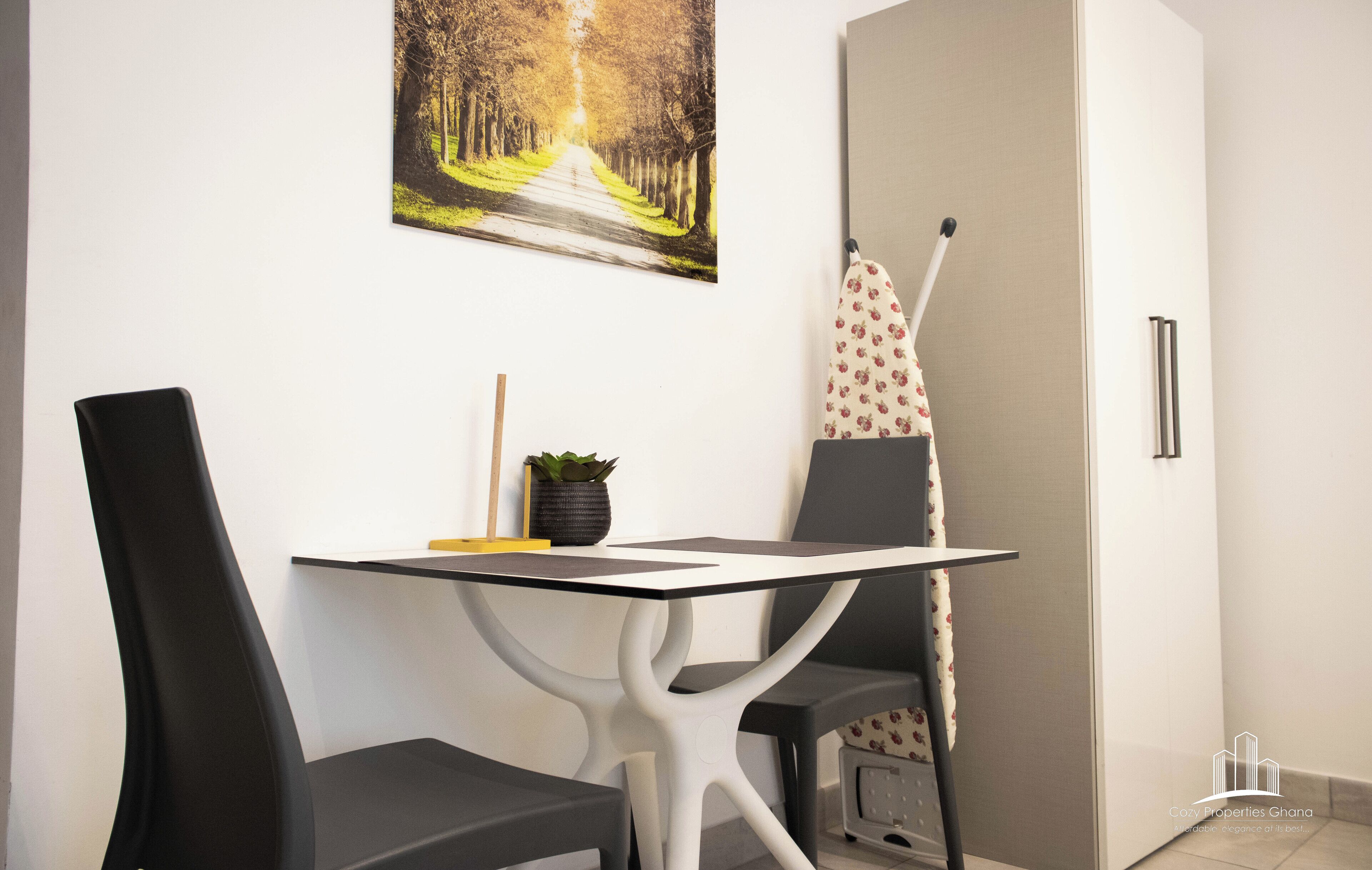 A modern room with a white table and chairs, a potted plant, and a painting of a tree-lined path on the wall.