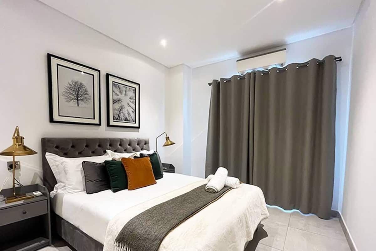 Un dormitorio con cabecera capitonada, cama con ropa blanca, dos cuadros enmarcados, una mesita de noche con lámpara y una ventana con cortinas.
