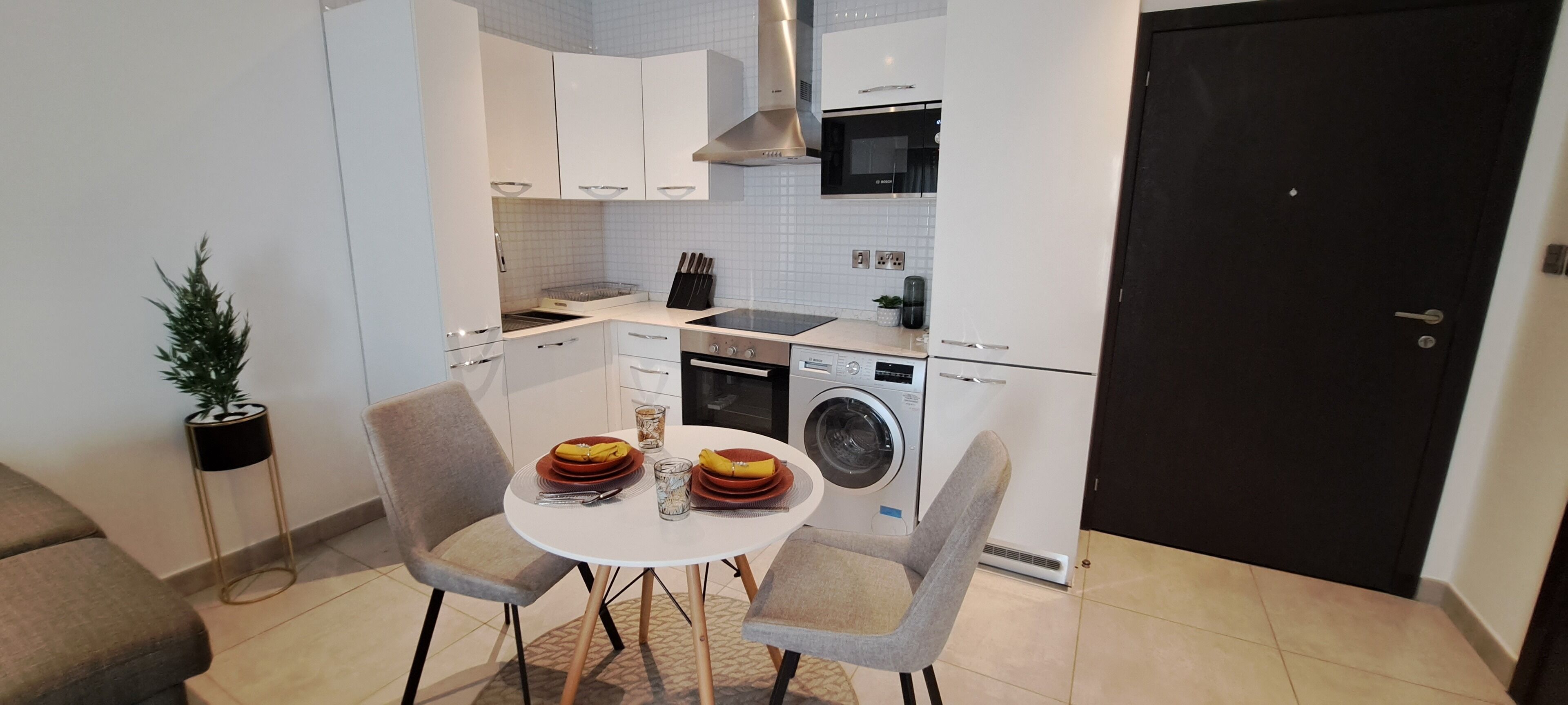 A compact kitchen with white cabinets, a microwave, a stove, and a washing machine. There is a dining table with chairs and a small potted plant.