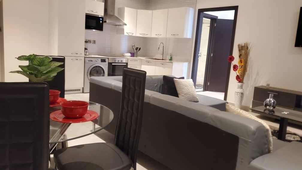 A modern kitchen with white cabinets, a built-in oven, and a washing machine. There is a dining area with a glass table and red placemats.