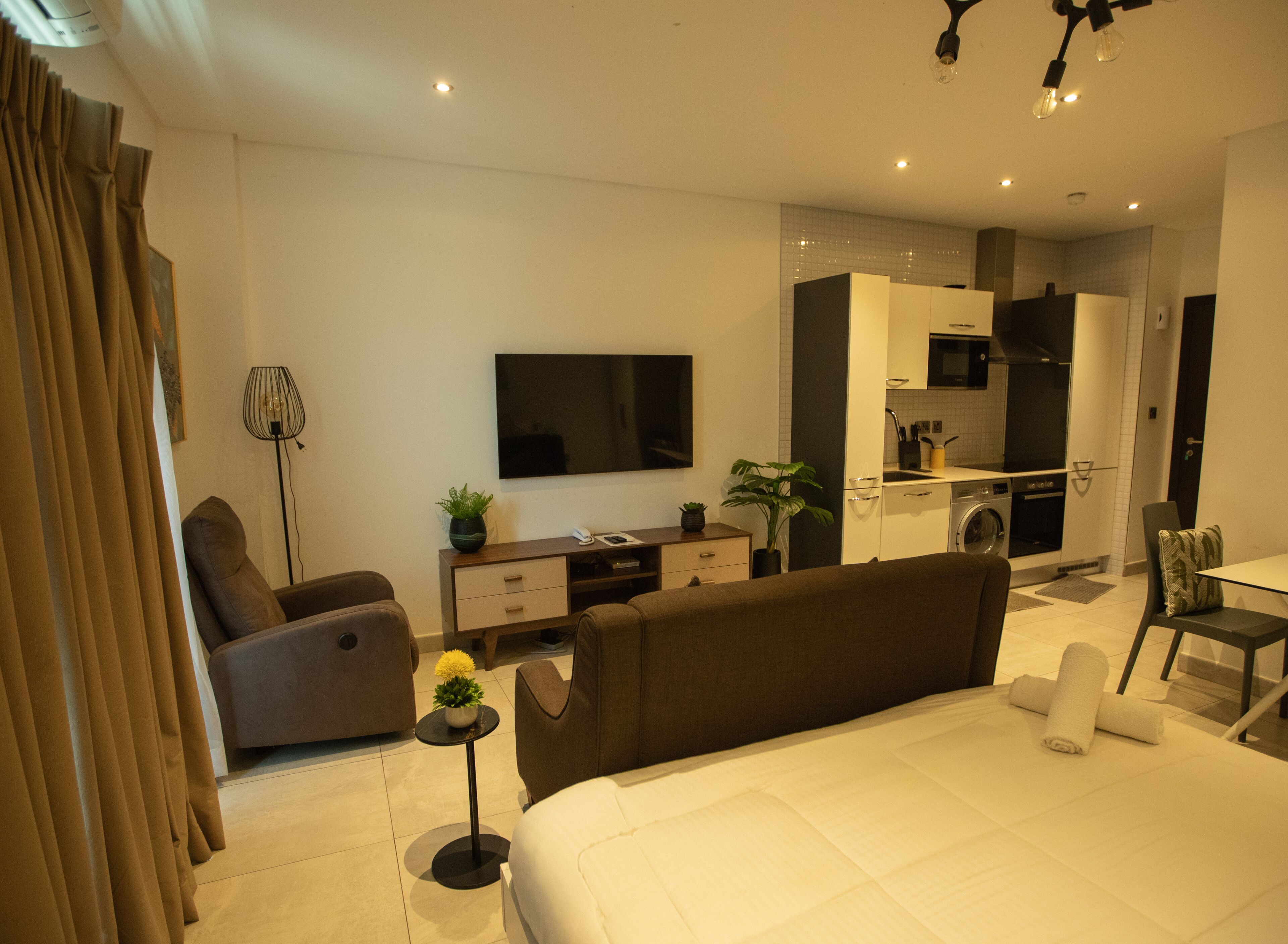 A modern hotel room with a large bed, a sofa, a TV, a small kitchen area, and a dining table.