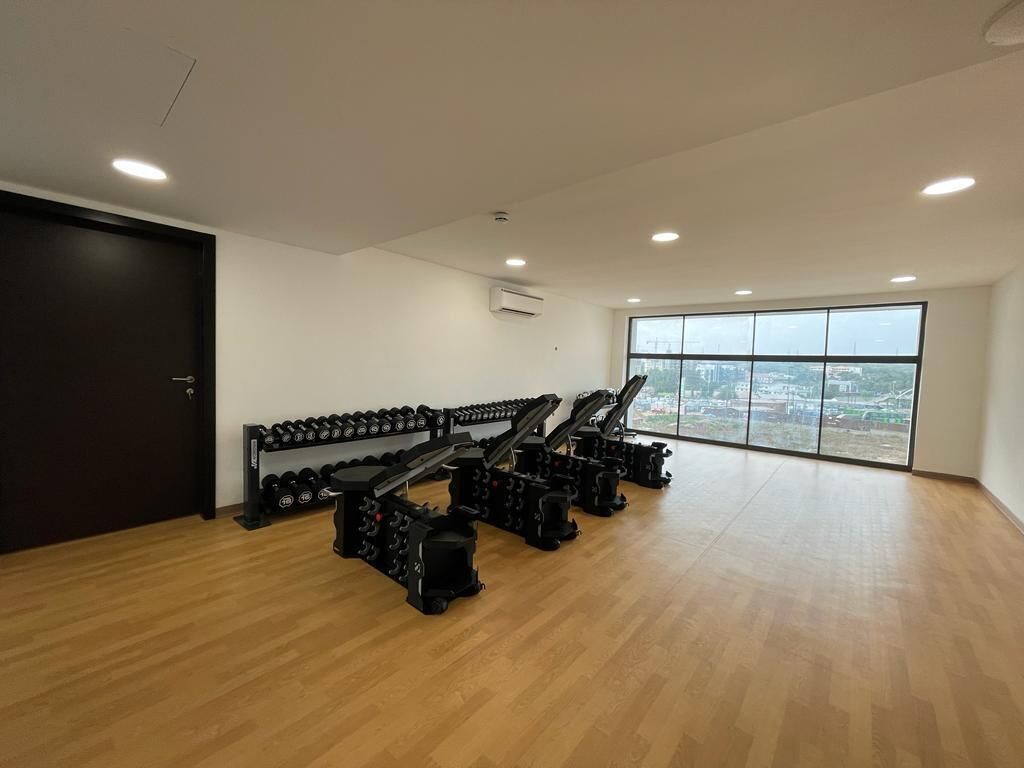 A gym with weightlifting equipment and dumbbells, large windows, and a view of the city.