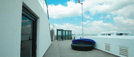 A rooftop terrace with a covered seating area, a glass door entrance, and a clear view of the sky and distant landscape.