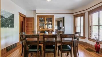 A dining area with a wooden table and chairs, a bookshelf, and a view through large windows.