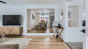 Ein modernes Wohnzimmer mit Fernseher, Holzmöbeln, einem Essbereich mit Chandelier und einer durch eine offene Tür einsehbarer Küche.